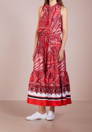 Femme portant une robe maxi sans manches rouge à motif cachemire avec ourlet à volants et baskets blanches, debout devant un fond neutre.