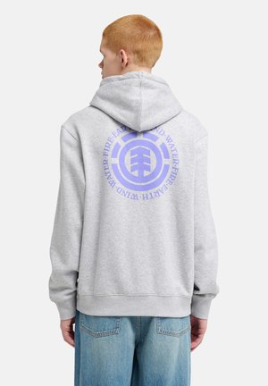 Jeune personne aux cheveux courts rouges portant un sweat à capuche gris clair avec un emblème circulaire bleu et un texte dans le dos, accompagné d’un jean bleu.