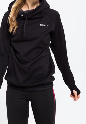 Kobieta w czarnej bluzie z kapturem z logo "CRAFT" i czarnych legginsach sportowych z różowym paskiem na białym tle.