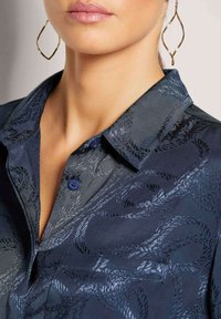 Marineblaue Bluse mit strukturiertem Design, ausgestattet mit einem Kragen, zwei blauen Knöpfen und einer Akzenttasche. Weicher Stoff mit dezentem Muster.