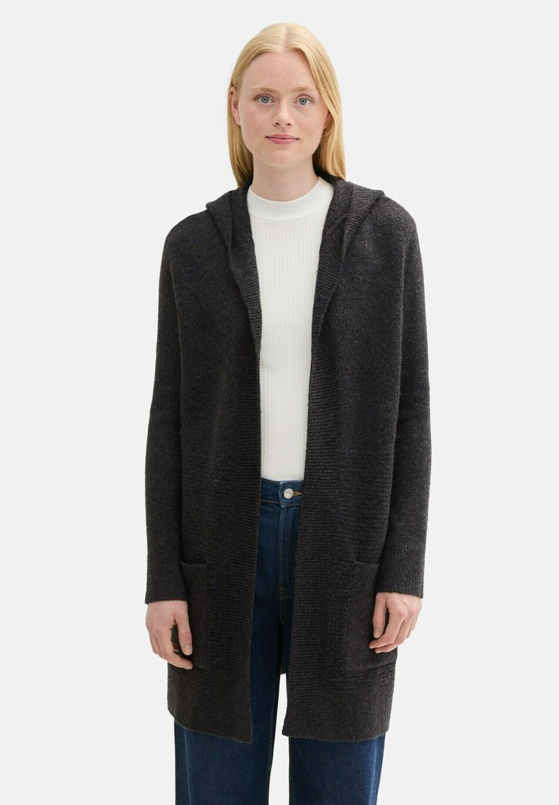 Mörkgrå huva cardigan gjord av ett texturerat stickat material med öppen fram, långa ärmar och två framfickor. Bärs över en vit topp och jeans.