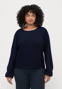 Pull en maille bleu marine avec un col rond, une texture côtelée et de longues manches légèrement évasées. Associé à un pantalon sombre.