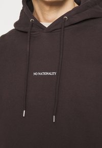 Ciemnobrązowa bluza z kapturem wykonana z miękkiego materiału, z przednią kieszenią kangurka, regulowanymi sznurkami oraz bia�łym napisem "NO NATIONALITY".