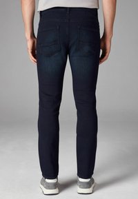 Vaqueros oscuros de corte slim-fit en índigo, con cinco bolsillos, un desgaste sutil y detalles de pespunte. Combinados con zapatillas grises y una camisa clara.