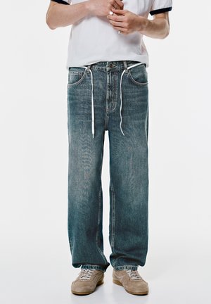 Hellblaue Jeans aus Denim mit entspannter Passform, ausgestattet mit weißen Kordeln, fünf Taschen und einem ausgewaschenen Look, kombiniert mit Schuhen in neutralen Farben.