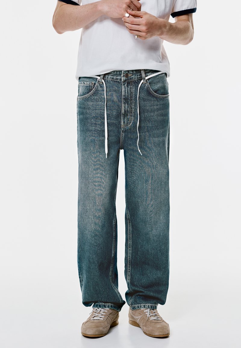 Hellblaue Jeans aus Denim mit entspannter Passform, ausgestattet mit weißen Kordeln, fünf Taschen und einem ausgewaschenen Look, kombiniert mit Schuhen in neutralen Farben.
