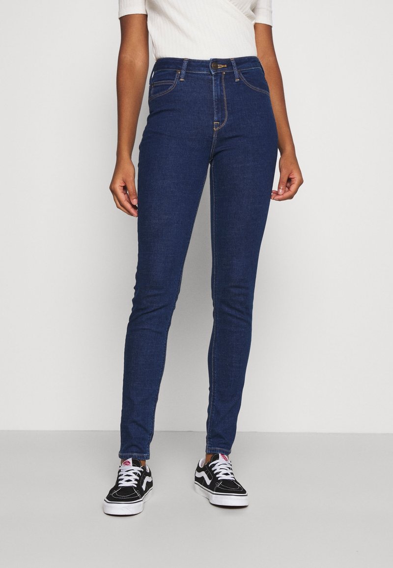 Höga, smala blå denimjeans med sex fickor, ton-i-ton sömmar och knäppning framtill, kombinerat med svarta sneakers.