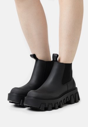 Bottes à plateforme chunky noires avec des tirettes, portées sur des jambes à la peau claire sur fond blanc.