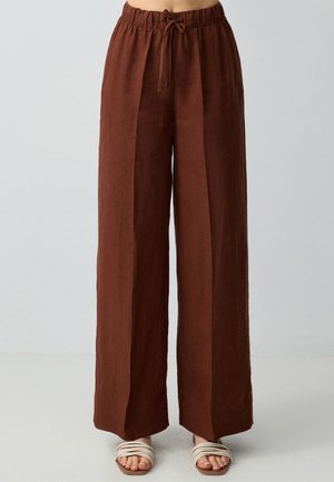 Pantalons large en lin marron avec une taille élastique et un cordon de serrage, dotés de coutures verticales et d'une finition texturée, associés à des sandales blanches.