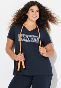 Marineblå v-hals t-shirt med hvid grafisk tekst, parret med mørke bukser. Modellen holder en orange sjippetov. Casual atletisk design og pasform.