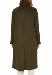 Osoba ubrana w długi, ciemnooliwkowy płaszcz wełniany widziana z tyłu, w połączeniu z białymi spodniami na jednolitym białym tle.