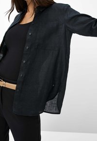 Chemise en lin noire avec fermeture boutonnée à l'avant, manches longues, poche poitrine et ourlet arrondi avec fentes latérales. Portée par-dessus un haut noir.