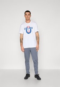 Hvid T-shirt med blå og rød "ENGLAND" grafik, parret med grå sweatpants med sidelæg, og sorte atletiske sko.