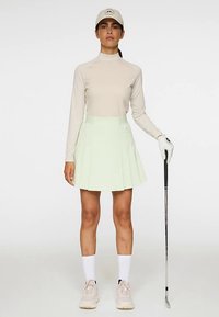 Donna con maglia beige a maniche lunghe, gonna plissettata verde chiaro, cappello bianco, guanti e scarpe, con in mano un bastone da golf, in piedi su sfondo bianco.