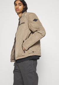 Tan quiltad jacka med dragkedja fram, sidofickor och en svart accentpatch på axeln. Tyget verkar slätt och lätt.