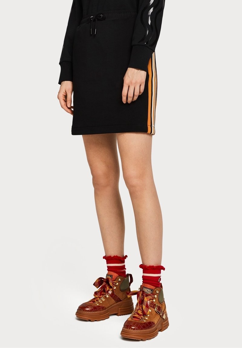 Zwarte sweatshirtjurk met zijstrepen in oranje; gecombineerd met stevige bruine laarzen met rode accenten en gestreepte rode sokken.