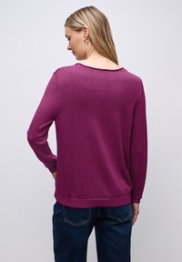 Lila lätt tröja med långa ärmar, rund halsringning och ribbat nederkant, kombinerad med mörkblå jeans, sedd bakifrån.