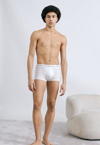 Λευκά ανδρικά boxer briefs με στενή ελαστική μέση. Το μοντέλο έχει αθλητική σωματική διάπλαση και στέκεται πάνω σε γκρι ανοιχτό δάπεδο, με μια κρεμ καρέκλα κοντά.