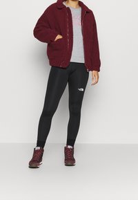 Giacca in pile bordeaux con zip frontale, t-shirt grigia sotto, leggings neri e scarponcini da trekking bordeaux con design con lacci e logo.