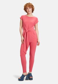 Lyserød jumpsuit med korte ærmer og knudedetalje i taljen, parret med blå denim platform sandaler. Glat tekstur, tætsiddende design.