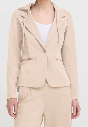 Beige skræddersyet blazer med hakkede revers, enkelt knaplukning og sidelommer, båret over en hvid T-shirt med rund hals og matchende beige bukser.
