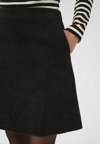 Čierna manšestrová sukňa s vertikálnou rebrovanou textúrou, vybavená bočnými vreckami a priliehavým pásom, v kombinácii s pruhovaným topom s dlhým rukávom.