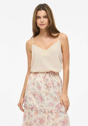 Tânără purtând un top bej fără mâneci și o fustă florală deschisă, cu flori roz și mov, stând pe un fundal alb.