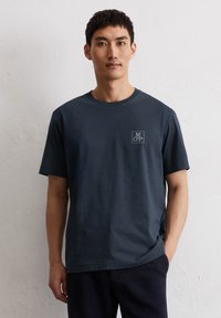 Dunkelblaues Baumwoll-T-Shirt mit Rundhalsausschnitt, kurzen Ärmeln und einem kleinen grauen Logo in einem Quadrat auf der linken Brust. Glatte Textur.