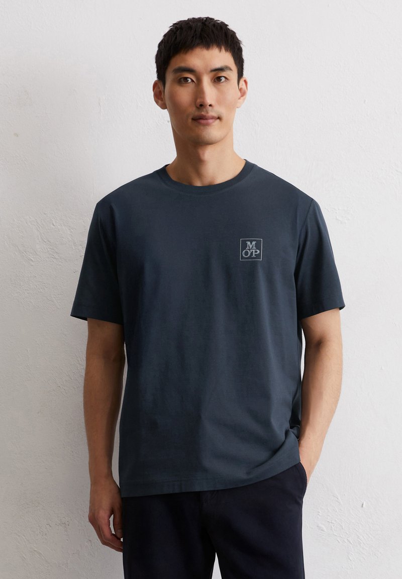 Dunkelblaues Baumwoll-T-Shirt mit Rundhalsausschnitt, kurzen Ärmeln und einem kleinen grauen Logo in einem Quadrat auf der linken Brust. Glatte Textur.