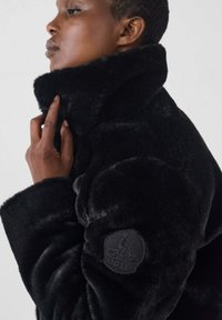 Cappotto in pelliccia sintetica nera con colletto alto, superficie testurizzata e una patch con logo sulla manica. Il design presenta una forma voluminosa e morbida.
