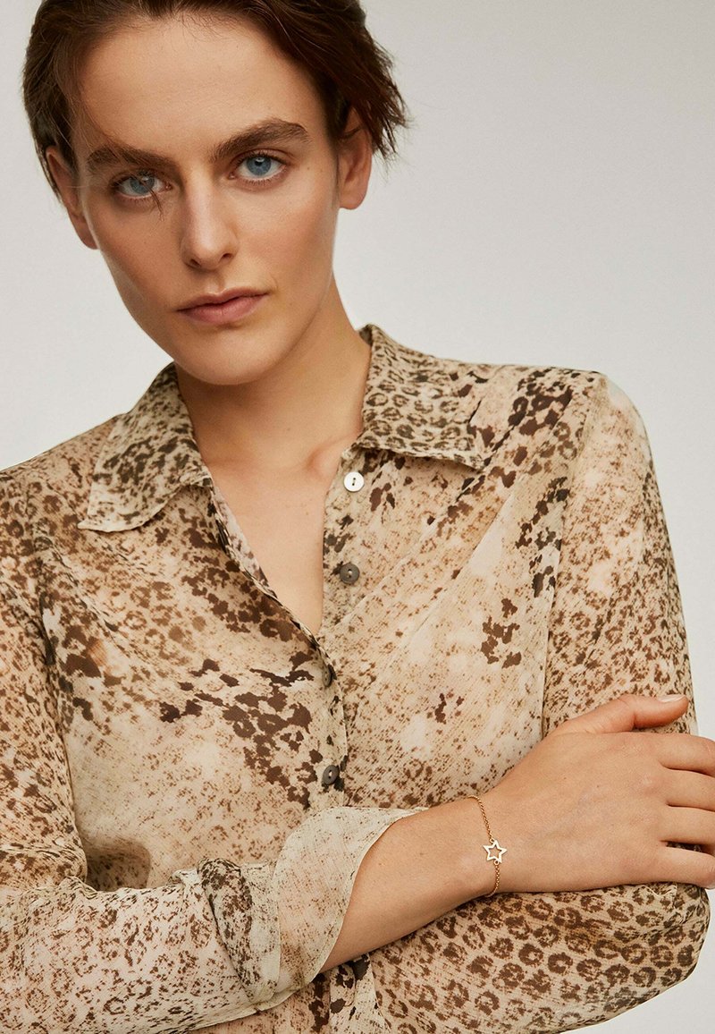 Blusa semitransparente con un patrón de estampado de leopardo en tonos cálidos de beige y marrón, con un cuello clásico y detalles de botones. Pulsera con estrellas doradas visible.