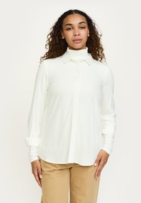 Camicia bianca a maniche lunghe con colletto classico e davanti con bottoni. Realizzata in tessuto leggero e liscio, con polsini risvoltati.