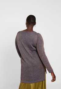 Kobieta o krótkich włosach ubrana w długi rękaw, błyszczącą fioletową bluzkę i oliwkowo-zieloną spódnicę, stoi odwrócona tyłem na szarym tle.