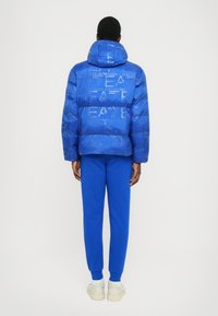 Chaqueta puffer azul con capucha que presenta un patrón de logo en relieve. Combinada con pantalones azules y zapatillas de color claro, vista desde atrás.