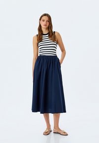 Top senza maniche a righe blu navy e bianco, abbinato a una gonna blu navy a tinta unita. La gonna presenta una vita arricciata e tasche laterali.