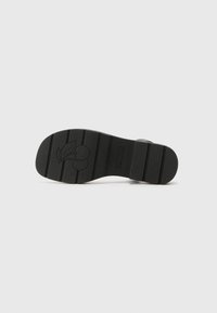 Semelle de chaussure en caoutchouc noir avec un motif texturé, présentant de profondes rainures et un logo cerise au talon. Design plat, finition sans couture.