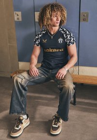 Marineblaues Trikot mit gemusterten Ärmeln, goldener Schrift und Logo; ausgebleichte hellblaue Jeans; braune und cremefarbene Sneakers mit schwarzen Akzenten.