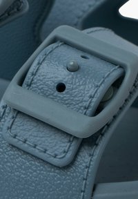 Lichtblauwe sandaalband van textuurleer en een rechthoekige plastic gesp. Onderdeel van contrasterende stiksel en een verstelbaar gatontwerp.