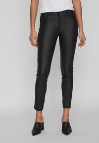 Pantalon skinny en simili cuir noir avec une taille haute, doté de poches avant et d'une texture lisse et brillante. Porté avec des mules à talons noires.