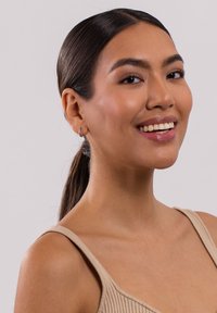 Porträt einer Person mit dunklem Haar im Pferdeschwanz, die ein geripptes beiges Oberteil und kleine silberne Creolen trägt. Glatte Haut und natürliches Make-up.