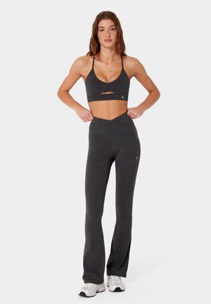 Frau trägt schwarzen Sport-BH mit Ausschnitt und schwarze Leggings mit hoher Taille und ausgestellten Beinenden, steht mit Händen in der Taille und weißen Sneakers.