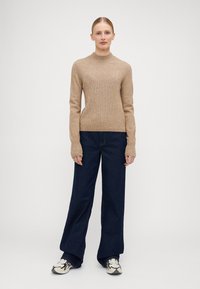 Beige, ribbet strikkegenser med høy hals og lange ermer. Kombinert med vide, mørk denim-bukser og mønstrede joggesko.