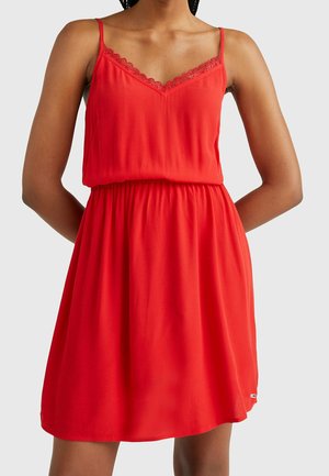 Femme portant une robe rouge vif sans manches avec un décolleté en V orné de dentelle et une taille élastique, les bras placés derrière le dos, sur un fond clair uni.