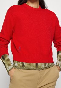 Henkilö, jolla on yllään kirkkaan punainen neulepaita vihertävän tahratut päällekkäinveto pitkähihainen paita, yhdistettynä vaaleisiin housuihin, kädet taskuissa.