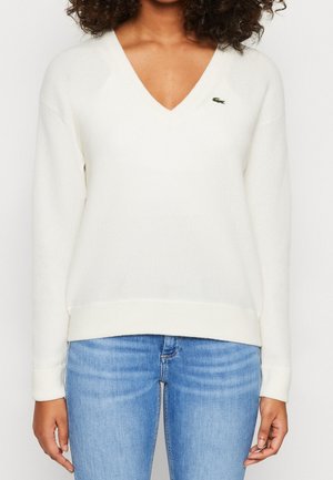 Femme portant un pull blanc à manches longues avec un col en V, orné d'un petit logo crocodile vert, et un jean bleu clair.