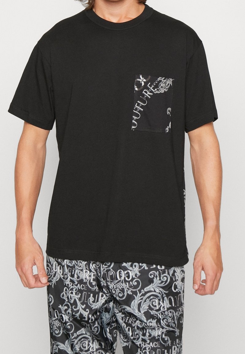 Man wearing black t-shirt with patterned pocket and black patterned pants featuring white swirling text and designs.