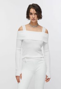 Haut blanc côtelé à épaules dénudées avec manches longues, orné de détails cloutés sur les bretelles. Porté avec un pantalon blanc ajusté.