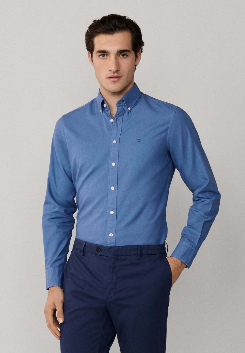 Junger Mann mit einem zugeknöpften blauen Hemd und dunkelblauer Hose, steht mit einer Hand in der Tasche vor einem schlichten hellen Hintergrund.