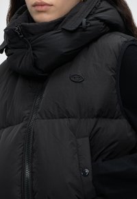Κοντινό πλάνο ενός ατόμου που φοράει μαύρο, καπιτονέ αμάνικο γιλέκο με ψηλό γιακά και φερμουάρ, πάνω από μαύρο μακρυμάνικο τοπ.