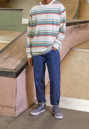 Personne portant une chemise blanche à manches longues avec des rayures horizontales colorées, un jean bleu et des baskets grises, debout près d'une rampe de skate.